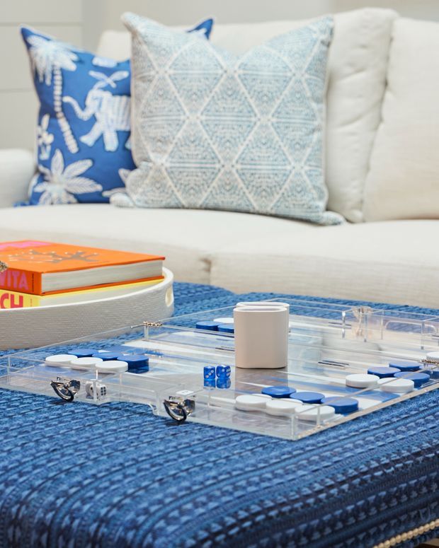 Close-up lifestyle view of a blue-textured upholstered ottoman or coffee table with a clear acrylic board game set up on top, including blue and white playing pieces, dice, and a small white cup. A white sofa with blue and pale patterned cushions sits in the background, creating a bright, casual living-room scene.