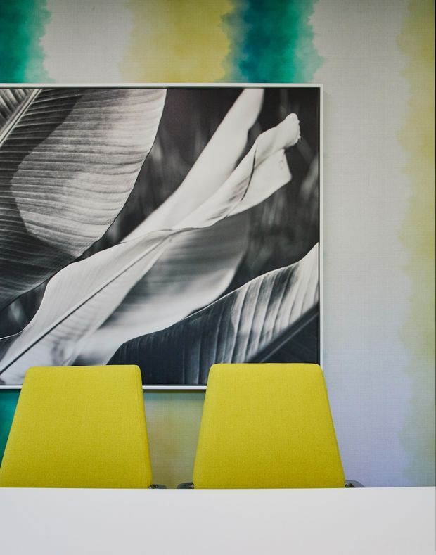 A contemporary interior vignette showing two bright lime-yellow upholstered dining or meeting chairs positioned behind a white tabletop. A large black-and-white botanical artwork hangs on the wall, which has a softly blended abstract mural in teal, yellow, grey, and pale tones.