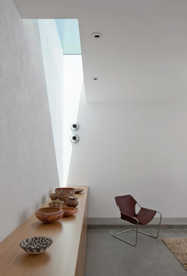 Minimalist interior with white textured plaster walls, a high angled skylight or clerestory opening, recessed ceiling lights, and a long light-wood display console holding ceramic bowls. A sculptural burgundy leather sling chair sits on a polished grey concrete floor, with a small neutral rug at the edge of the frame.