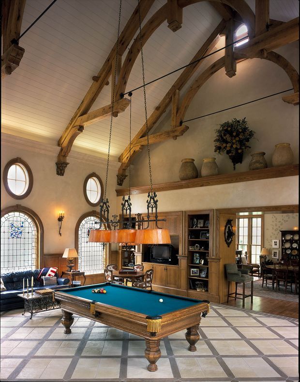 A grand traditional games room centred on a full-size billiards table, with exposed heavy timber trusses, a vaulted white-painted ceiling, arched leaded-glass windows, oak built-in cabinetry, decorative shelving, and a large rustic chandelier. A sitting area and dining area are visible beyond the open doorway. The room has a warm, refined, heritage-inspired character.