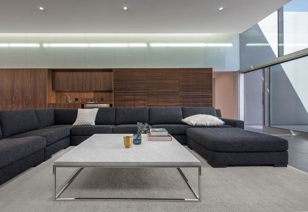 A spacious contemporary living room with a large dark charcoal sectional sofa, oversized light stone coffee table, and pale textured rug. The room features warm dark timber built-in cabinetry and a slatted timber wall, recessed ceiling lights, linear concealed lighting, and extensive floor-to-ceiling glazing with strong geometric exterior shadows. The palette is restrained and refined, combining grey, white, and dark wood tones.