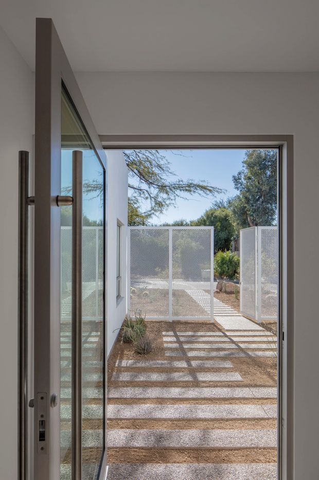 View from inside through an open glazed door toward a landscaped courtyard or side passage. The minimalist white interior opening frames a gravel and concrete stepping-stone path, with perforated white metal privacy screens and a simple planted garden beyond. Strong sunlight creates patterned shadows across the path.