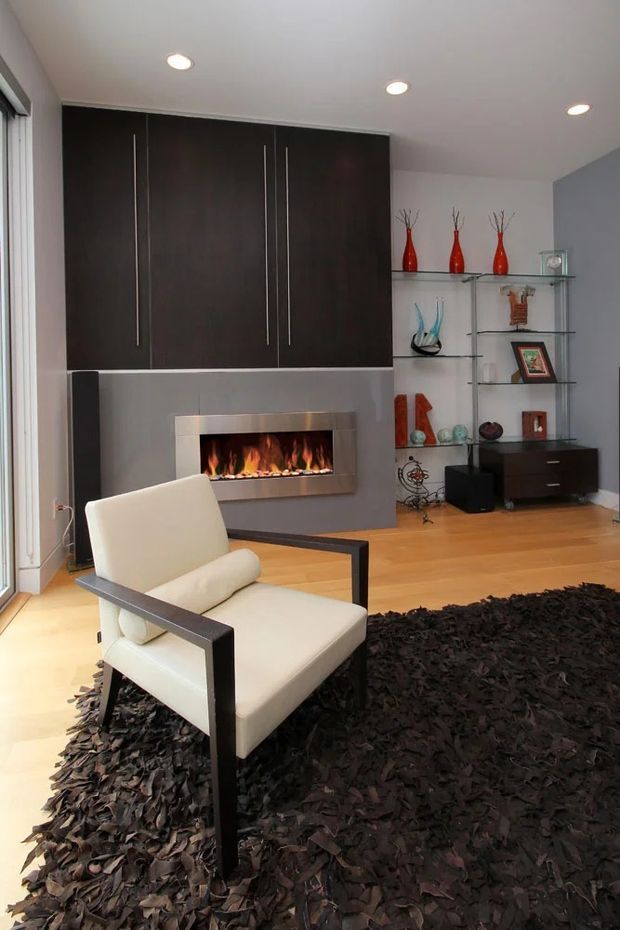 Contemporary living room with a dramatic dark built-in entertainment and fireplace wall, glass display shelving, timber flooring, recessed ceiling lights, and a white upholstered armchair over a large shaggy dark rug. A linear fireplace forms the focal point beneath the dark cabinetry, with decorative objects arranged on open shelving.