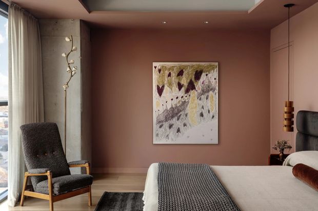 A warm, refined bedroom with dusty pink walls, a concrete feature column, full-height curtains and glazing, a dark upholstered lounge chair, a neatly made bed, a sculptural pendant light, and abstract artwork. The room combines soft textiles and timber details with visible concrete and contemporary furnishings.