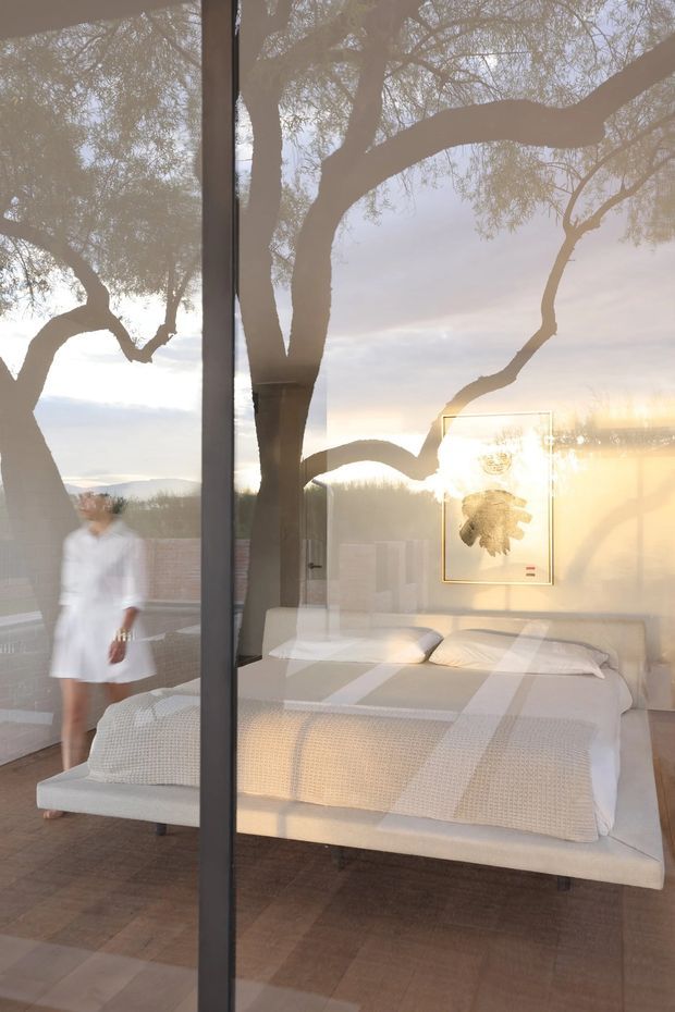 A serene bedroom viewed through floor-to-ceiling glazing, with a neatly made bed, white textured bedding, a pale upholstered bed base, and a framed botanical artwork. The glass strongly reflects large trees and the sunset landscape outside, creating layered indoor-outdoor imagery. A blurred person is visible near the left side, and the room has warm, soft natural light with dark timber flooring and a minimal, refined appearance.