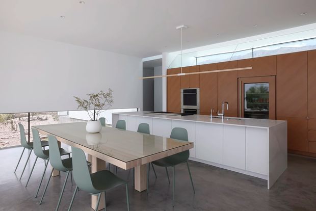 A bright, open-plan contemporary kitchen and dining area with a long pale stone dining table, muted green dining chairs, and a large white island. The kitchen features full-height warm timber cabinetry, integrated appliances, a sink, minimalist pendant lighting, polished concrete flooring, and expansive glazing with a low landscape view beyond.