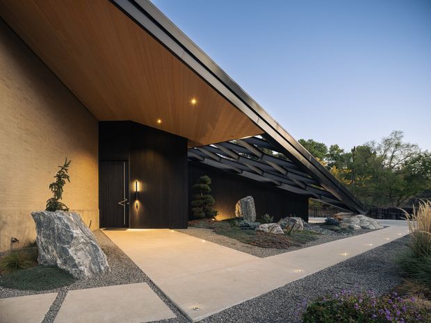 Exterior architectural view of a refined contemporary single-storey structure with a dramatic projecting roof and deep timber-lined soffit. A dark entrance volume sits beneath the canopy, while layered black structural elements extend over a landscaped gravel garden with sculptural rocks, clipped trees and ornamental planting. Concrete paths lead through the minimalist courtyard-like approach.