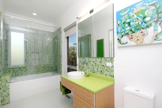 Bright contemporary bathroom with a bathtub enclosed in a green and white mosaic-tiled shower area, a floating timber vanity with a vivid lime-green benchtop, round vessel basin, large mirrored cabinet, and a white toilet. A colourful abstract artwork adds a playful accent.
