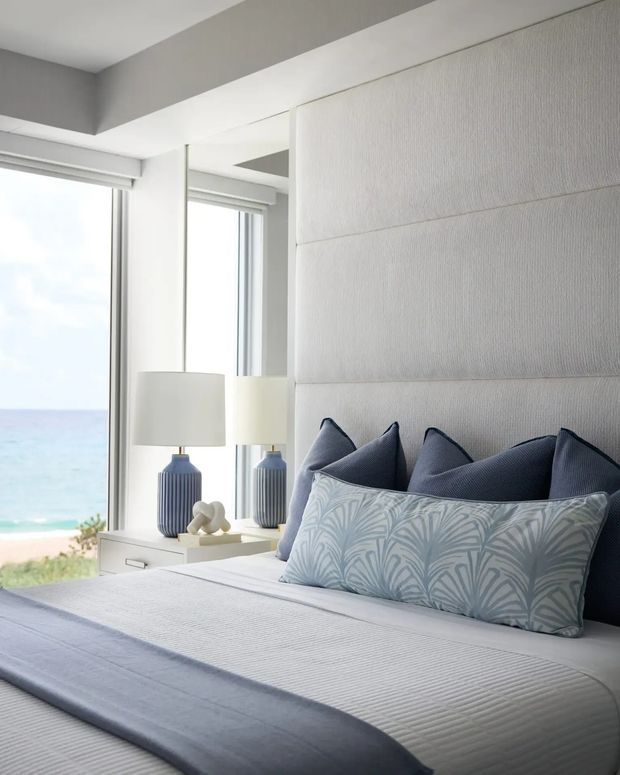Bright coastal bedroom with a neatly made bed positioned beside floor-to-ceiling windows overlooking the beach and ocean. The room features a padded, horizontally panelled light wall behind the bed, mirrored bedside cabinetry, blue-grey decorative pillows, patterned accent cushions, ceramic lamps, and a soft neutral palette.