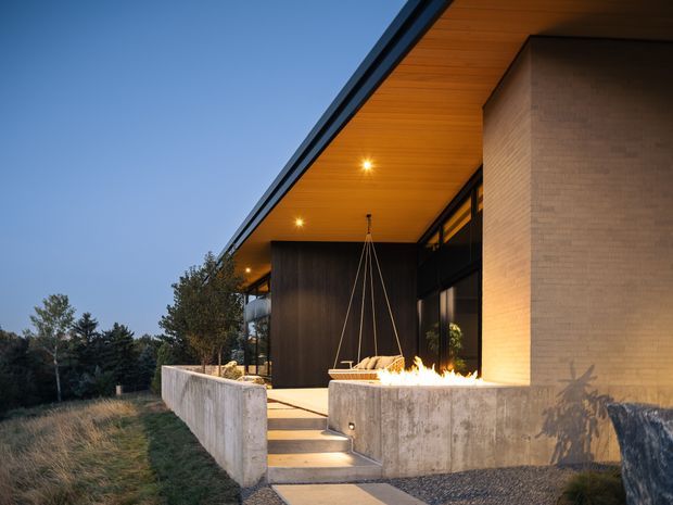 Exterior architectural photograph at dusk showing a modern covered terrace or entry walkway. The building has a broad flat projecting roof with warm timber soffit, dark metal-framed glazing, a large pale brick wall, exposed concrete retaining walls, and a linear outdoor fire feature beside the steps. Natural landscaping surrounds the structure.