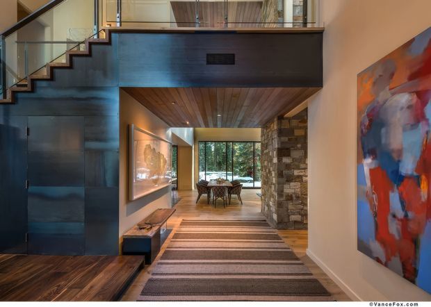 A sophisticated open-plan residential interior viewed from an entry hall, featuring a floating industrial-style staircase and dark metal mezzanine structure, warm timber flooring and ceiling, a textured natural stone wall, large glazing, and a dining area beyond. A colourful abstract artwork adds a bold accent to the otherwise warm, refined palette.