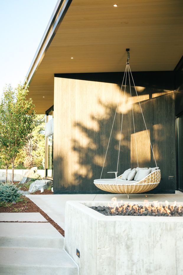 Contemporary covered outdoor terrace with a timber-lined soffit, dark vertical wall cladding, expansive glazing, and a suspended woven hanging chair. A concrete fire feature with visible flames forms the foreground, while pale concrete paving and landscaped planting frame the space.