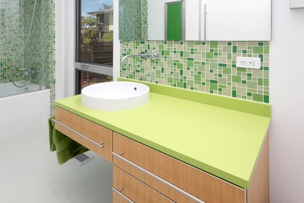 Bright contemporary bathroom featuring a lime-green vanity benchtop with integrated raised round basin, warm timber-look drawer cabinetry, a green mosaic tile splashback, white wall cabinetry, and a white bathtub with green mosaic wall tiles. A window provides natural light.