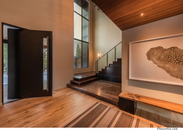 A warm, contemporary entry foyer with a tall dark timber front door, expansive vertical glazing, a compact staircase with dark timber steps and glass balustrade, and a richly timber-lined ceiling. The space features timber flooring, a built-in timber console bench, recessed lighting, and a large framed organic wall artwork.