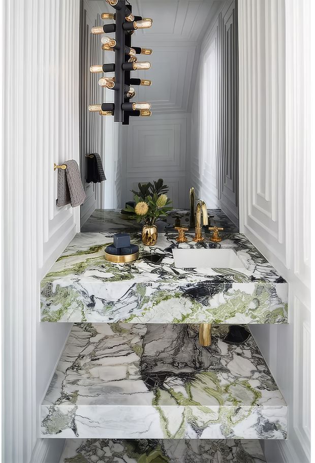 A narrow, highly decorative powder room with white raised wall panelling, a mirrored rear wall, and dramatic green, grey, black and white veined marble used across the floating vanity, benchtop, and lower wall or floor surfaces. The compact vanity includes a white vessel-style basin, sculptural gold tapware, matching gold hardware, a small floral arrangement, and a striking black pendant light with exposed warm bulbs.