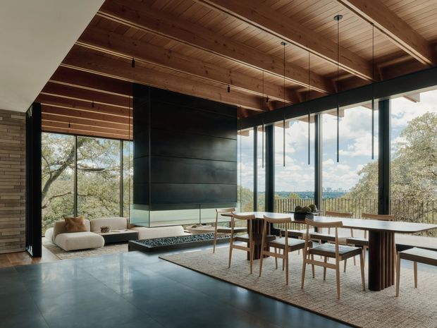 Open-plan contemporary living and dining area with a dramatic double-height timber-lined ceiling, dark stone or metal feature fireplace wall, expansive black-framed glazing, and views to trees and a distant urban landscape. A dining table occupies the foreground, while a low lounge seating area and linear fireplace sit beyond.