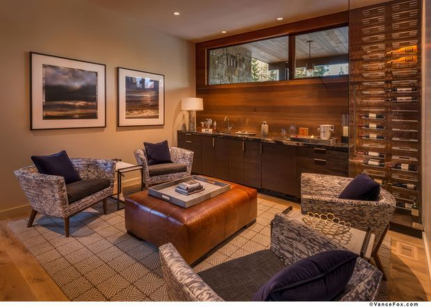 A warm, richly detailed lounge and home bar area with four patterned upholstered armchairs arranged around a large brown leather ottoman and low tray table. The room features dark timber built-in cabinetry, a stone or tile backsplash, an integrated bar counter, extensive wine storage, framed artwork, recessed ceiling lights, and a large interior window opening to another space. A geometric rug covers much of the timber floor.