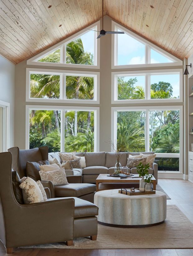 Bright, spacious living room with a dramatic vaulted timber-lined ceiling, expansive white-framed geometric windows, and lush tropical greenery visible outside. The room is furnished with a large neutral sectional sofa, leather armchairs, layered cushions, a textured area rug, wood coffee table, and a round upholstered ottoman. A ceiling fan and built-in shelving complete the relaxed, refined interior.