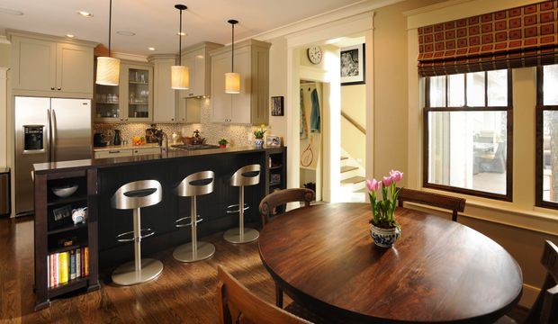 Warm, traditionally styled open-plan kitchen and dining area. The kitchen features cream raised-panel cabinetry, stainless-steel appliances, a dark island with four bar stools, glass-front storage, and pendant lighting. A large round dark timber dining table occupies the foreground beside a window with patterned Roman blinds. Timber flooring, decorative mouldings, and an adjacent stairway are visible.