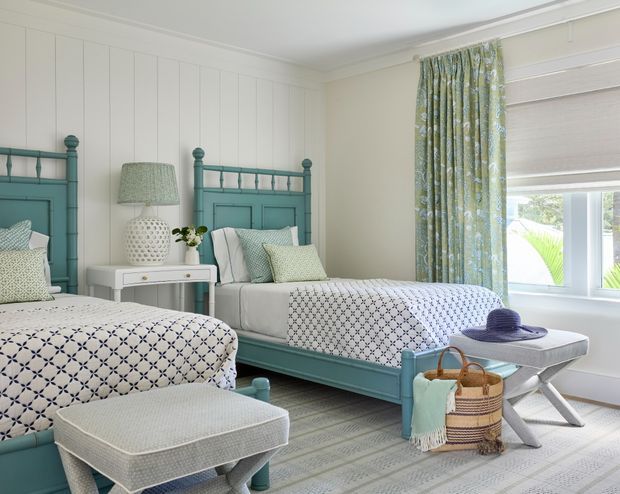 Bright, inviting twin bedroom with two teal-painted beds, patterned white-and-blue bedding, a matching upholstered bench, white vertical wall panelling, a small bedside table, decorative lamp, and floral curtains beside a large window. The room has a fresh coastal-inspired palette with soft green, teal, white, and light grey tones.