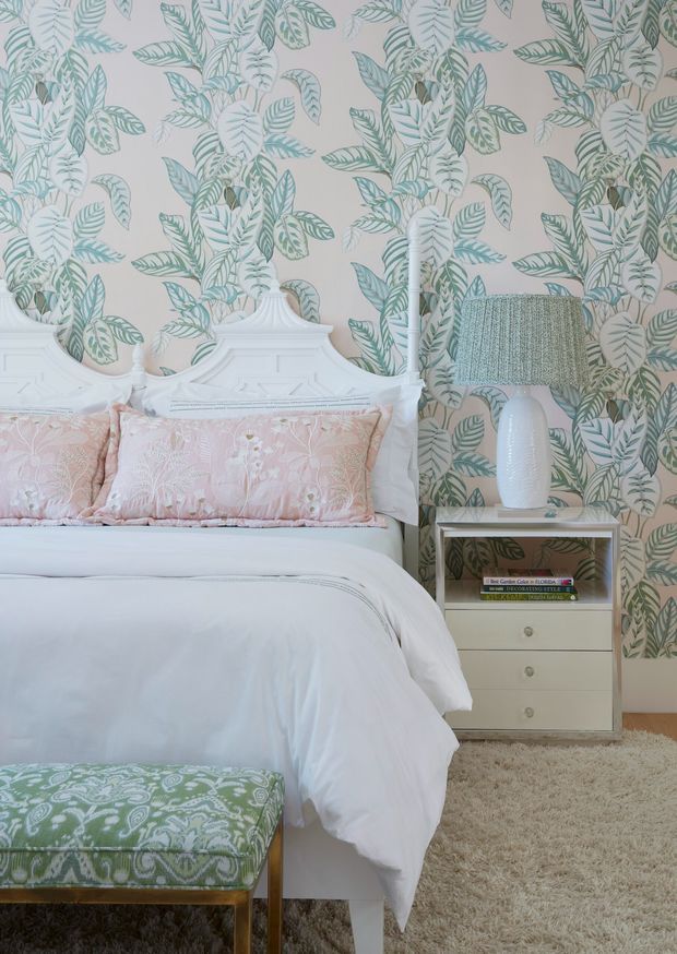 A softly styled bedroom featuring a white ornate bedhead, white bedding, blush-pink embroidered cushions, and a green patterned upholstered bench. The walls are covered in a pale botanical leaf wallpaper in sage, teal, and muted green tones. A small cream bedside table with drawers supports a white ceramic lamp with a textured pale green shade. A plush beige carpet adds warmth underfoot.