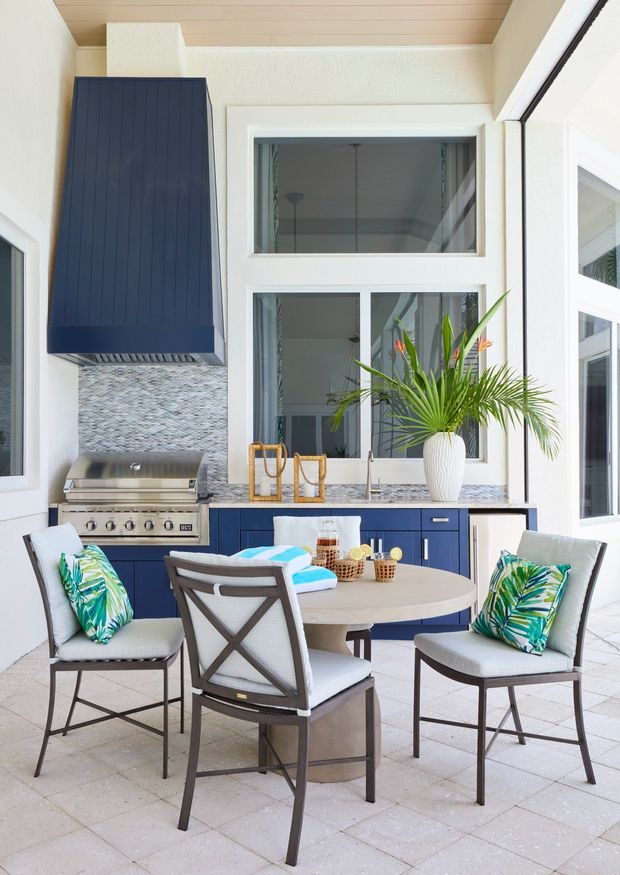 Bright covered outdoor dining and kitchen area with a built-in barbecue, dark navy cabinetry, textured light stone backsplash, pale tiled floor, and large white-framed windows. A round dining table with four chairs sits in the foreground, with tropical-patterned cushions and a potted palm adding a coastal resort feel.