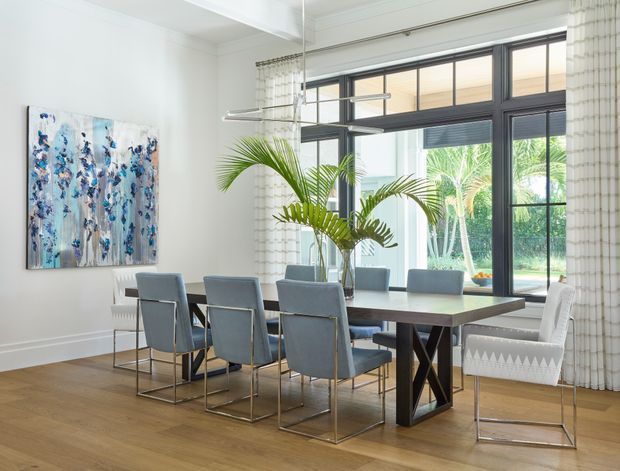 Bright, refined dining room with a large rectangular timber dining table surrounded by upholstered grey chairs. The room features warm timber flooring, white walls and curtains, expansive dark-framed windows overlooking greenery, a contemporary linear pendant light, tropical palm arrangement, and abstract blue-toned artwork.