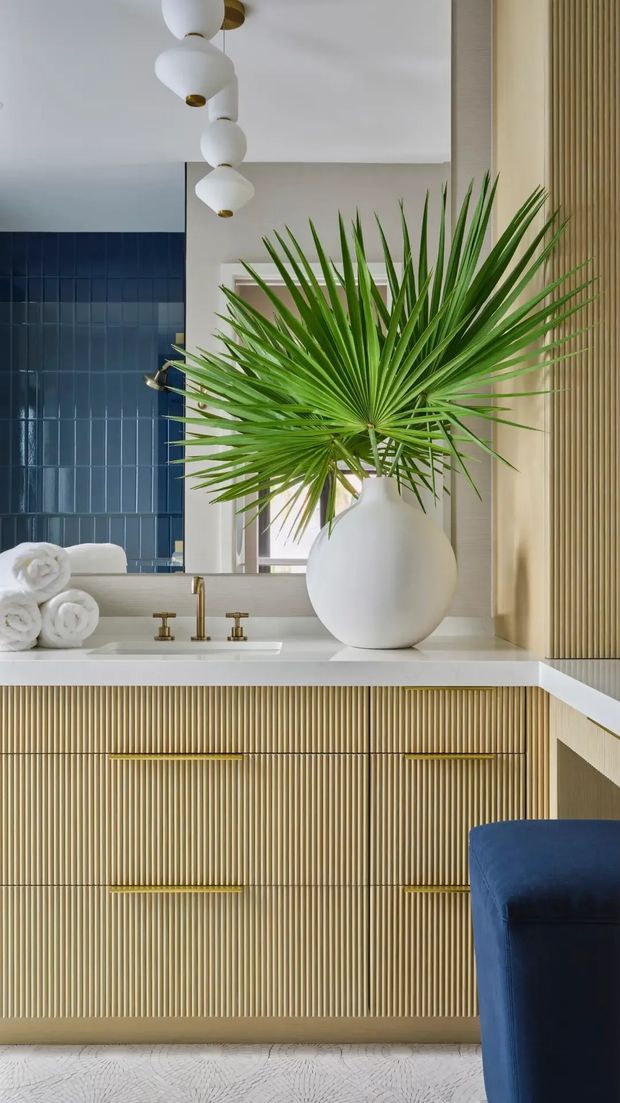 A refined bathroom vanity vignette featuring fluted champagne-gold cabinetry, a white countertop with brass tapware, a large round white vessel, and a lush fan palm. Deep blue glazed wall tiles contrast with pale walls, vertical timber detailing, and a plush blue upholstered element. A sculptural cluster pendant hangs overhead, creating a luxe, contemporary atmosphere.