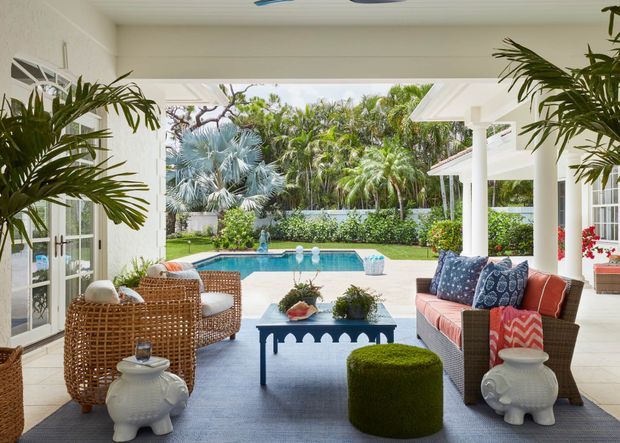 A bright, covered outdoor living area opening onto a landscaped garden and rectangular swimming pool. The space features wicker lounge chairs, a woven sofa with colourful cushions, a blue coffee table, tiled flooring, white columns and ceiling, and tropical planting beyond the pool.