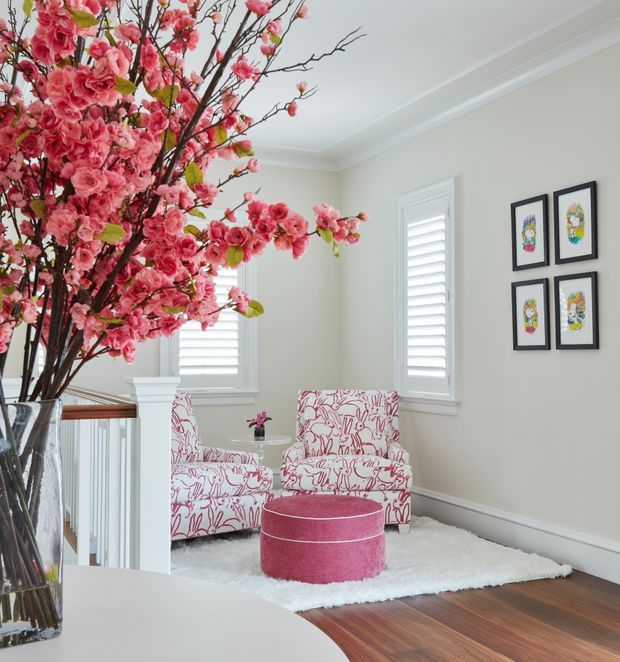 Bright, refined upstairs sitting nook or landing with pale walls, decorative ceiling cornices, plantation shutters, dark timber flooring, and a white plush rug. Two upholstered armchairs and a round pink ottoman create a small seating area, while framed colourful artwork decorates the wall. A large arrangement of pink blossom branches in a glass vase forms a prominent decorative feature.