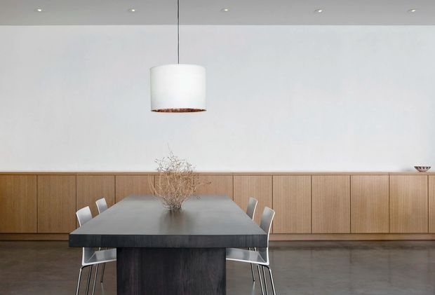 Minimal dining room with a long dark dining table and contemporary chairs, set against a full-width wall of warm timber built-in cabinetry. The room has a polished grey concrete floor, plain white walls, recessed ceiling lights, and a large suspended white pendant light with a timber-toned underside. The restrained composition and natural materials create a calm, refined contemporary interior.