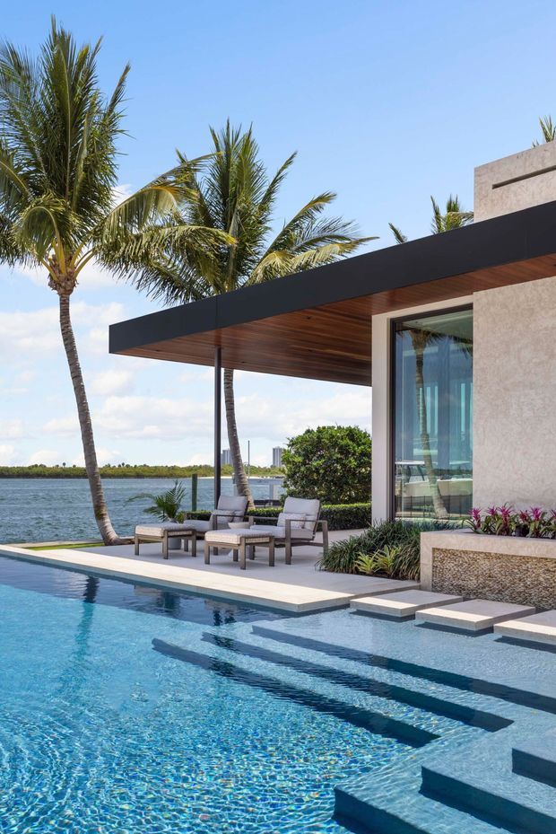 A modern waterfront residence with a large turquoise swimming pool in the foreground, broad submerged entry steps, and a raised pool deck furnished with outdoor lounge chairs. The house has a flat roof with a deep cantilevered overhang, warm timber soffit, dark fascia, expansive glazing, and textured light-coloured walls. Palm trees, landscaped planting, and a calm body of water reinforce the tropical coastal setting.
