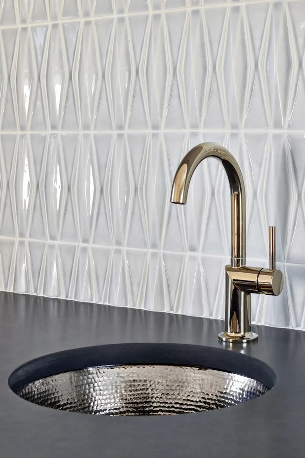 Close-up of a refined bathroom vanity featuring a hammered silver metal vessel sink set into a dark charcoal benchtop, a polished brass/gold curved mixer tap, and glossy white dimensional textured wall tiles with elongated linear relief.