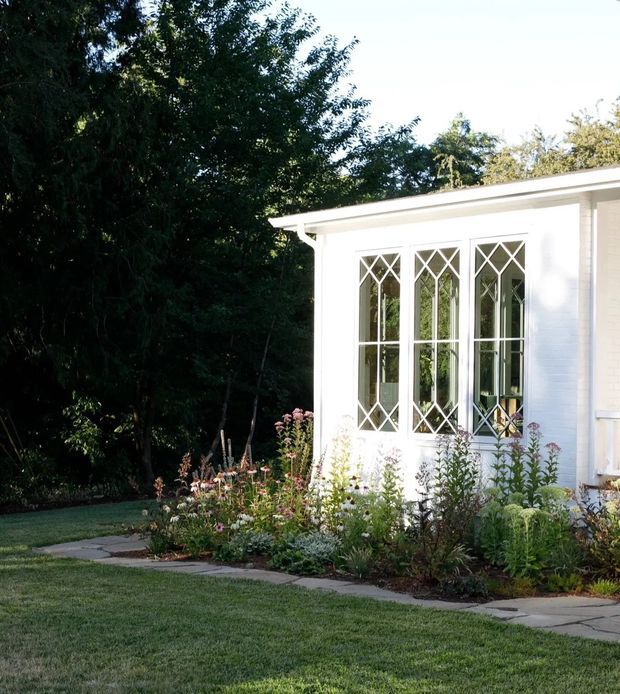 Exterior view of a white-painted brick garden room or house extension, featuring tall multi-pane windows with prominent diamond-pattern muntins and a shallow projecting roof. A planted garden bed, paved path, lawn, and mature trees surround the building.
