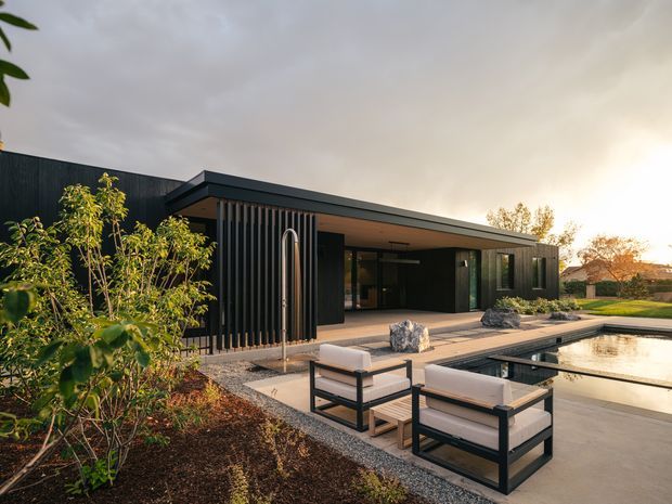 Contemporary single-storey residence with a dark vertical-clad facade and broad low-pitched roof extending over a covered outdoor entertaining area. The image shows a landscaped garden, paved terrace with modern outdoor lounge chairs, and a reflecting or swimming pool beside the house. Large glazed openings connect the interior to the terrace, while warm low-angle light and natural planting soften the dark architectural palette.