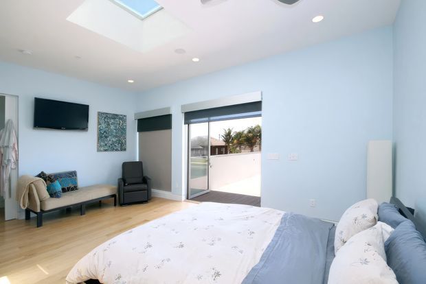 Bright, airy bedroom with pale blue walls, light timber flooring, a large bed with blue-and-white bedding, a small seating area, wall-mounted television, roller blinds, and wide glazed doors opening to an outdoor terrace. A skylight and recessed ceiling lights provide abundant natural and artificial light.