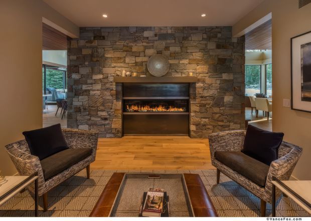 Warm, refined living room centered on a substantial stacked-stone fireplace wall with an inset linear fire. Two patterned armchairs with dark cushions face a low rectangular coffee table on a geometric area rug, while timber flooring and recessed ceiling lights add warmth. Dining and exterior views are visible through side openings.