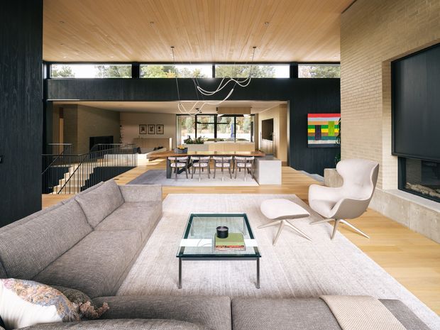 Open-plan contemporary living area with a large grey sectional sofa, glass coffee table, sculptural lounge chair and ottoman, and a fireplace with wall-mounted television. The space opens directly toward a dining area and kitchen, with a dramatic double-height volume, upper clerestory windows, dark feature walls, pale timber flooring, and a warm timber-lined ceiling.