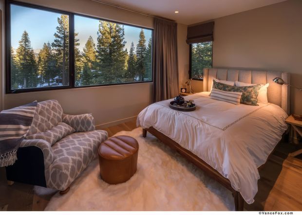 A warm, softly lit bedroom with a large upholstered bed positioned beside expansive windows overlooking tall evergreen trees. The room includes a patterned armchair, a small round leather ottoman, bedside tables, layered bedding, timber flooring, full-height curtains, and a plush light-coloured rug. The atmosphere is cozy and refined, with a strong connection to the wooded setting outside.