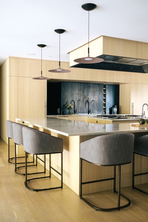 A bright contemporary kitchen centered on a large island with integrated seating. The space features extensive light timber cabinetry, a pale stone island benchtop, dark metal-framed upholstered bar stools, a dark metallic splashback, and a concealed rangehood with dark underside. Several slim pendant lights hang above the island, creating a refined, warm-toned interior.
