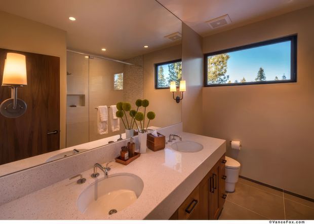 A warm, contemporary bathroom featuring a long double vanity with pale engineered-stone countertop, integrated oval basins, timber cabinetry, wall-mounted mirrors, a glass-enclosed shower, and a freestanding toilet. Recessed ceiling lights, decorative wall sconces, and a horizontal picture window provide illumination, with trees visible outside.