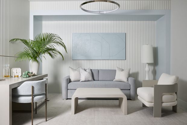 A refined, light-toned living room with vertically fluted wall panelling, a grey sofa, sculptural white armchairs, a rounded rectangular coffee table, a large potted palm, and a slim circular pendant light. The space has a calm, contemporary appearance with soft neutral furnishings and built-in or integrated joinery along the left side.