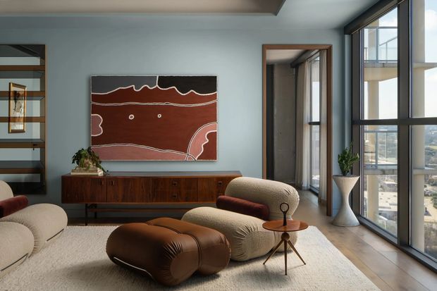 A refined contemporary living room in a high-rise setting, featuring floor-to-ceiling windows, a warm timber sideboard, sculptural upholstered lounge chairs, a brown leather ottoman, and a large abstract artwork. The room has pale blue-grey walls, a light timber floor, and abundant natural light with an urban view beyond the glazing.