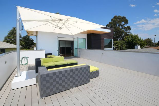 A bright rooftop terrace with pale timber-look decking, low white parapet walls and a large white cantilevered shade canopy. Contemporary outdoor seating in lime green and charcoal occupies the foreground, with sliding glass doors opening into a modern upper-level room and dark horizontal wall cladding behind.