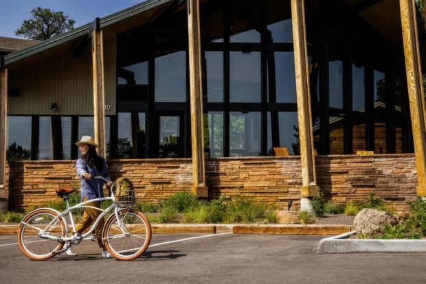 Exterior view of a lodge-like building with tall dark-framed windows, timber posts, a low-pitched roof, vertical wood cladding, and a stacked stone lower facade. A paved driveway and bicycle with a person are in the foreground, with trees visible in the background.
