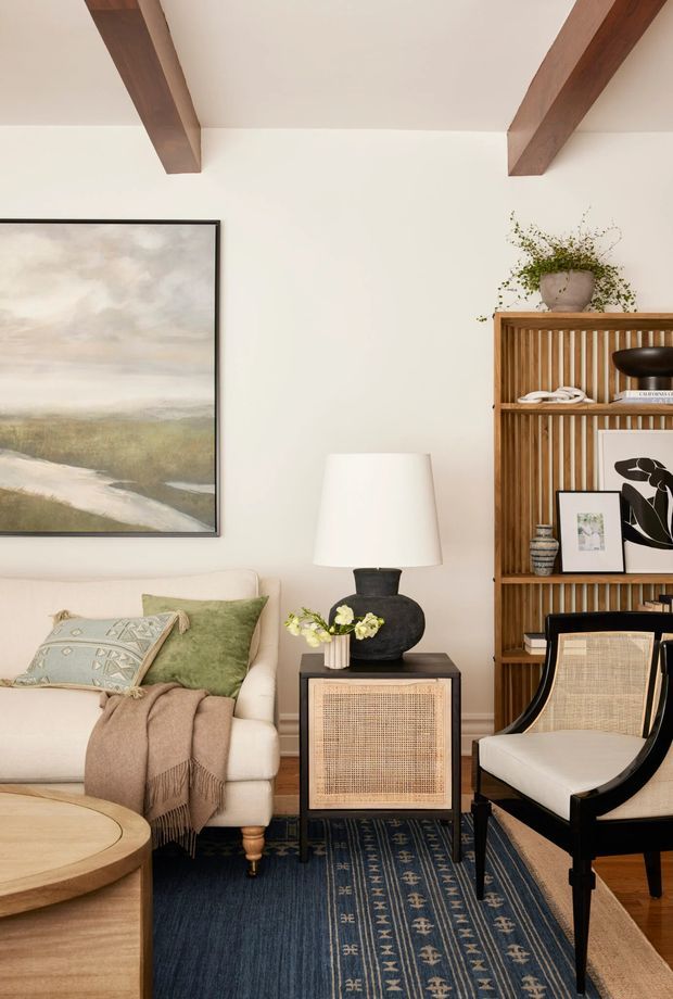 A warm, refined living room vignette with exposed dark timber ceiling beams, creamy plaster walls, a pale upholstered sofa, patterned cushions, a draped throw, a round timber coffee table, and a dark side table with woven cane-front cabinetry. A tall timber shelving unit with vertical slats displays books, artwork, ceramics, and greenery. A black-framed lounge chair, traditional landscape artwork, layered rugs, and a sculptural table lamp complete the room.
