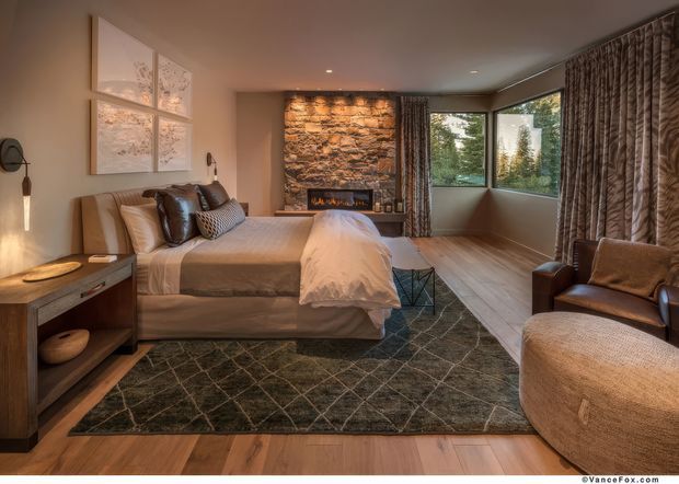 Warm, refined bedroom with a large upholstered bed, layered neutral bedding, leather and patterned pillows, timber flooring, a dark geometric rug, and a sculptural upholstered armchair. A dramatic exposed natural-stone feature wall incorporates a fireplace, while floor-to-ceiling patterned curtains and corner glazing bring in views of greenery. Contemporary lighting and timber bedside joinery complete the inviting interior.