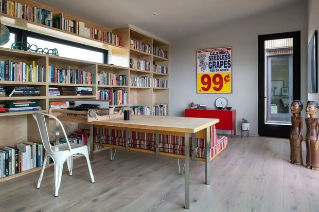 A bright, eclectic home office and reading room with extensive floor-to-ceiling built-in bookshelves, a large timber worktable, metal chairs, and a patterned upholstered bench. A vintage-style California seedless grapes poster adds a bold red and yellow accent, while a doorway leads to another interior space. The room has pale timber flooring, white walls, and abundant natural light.