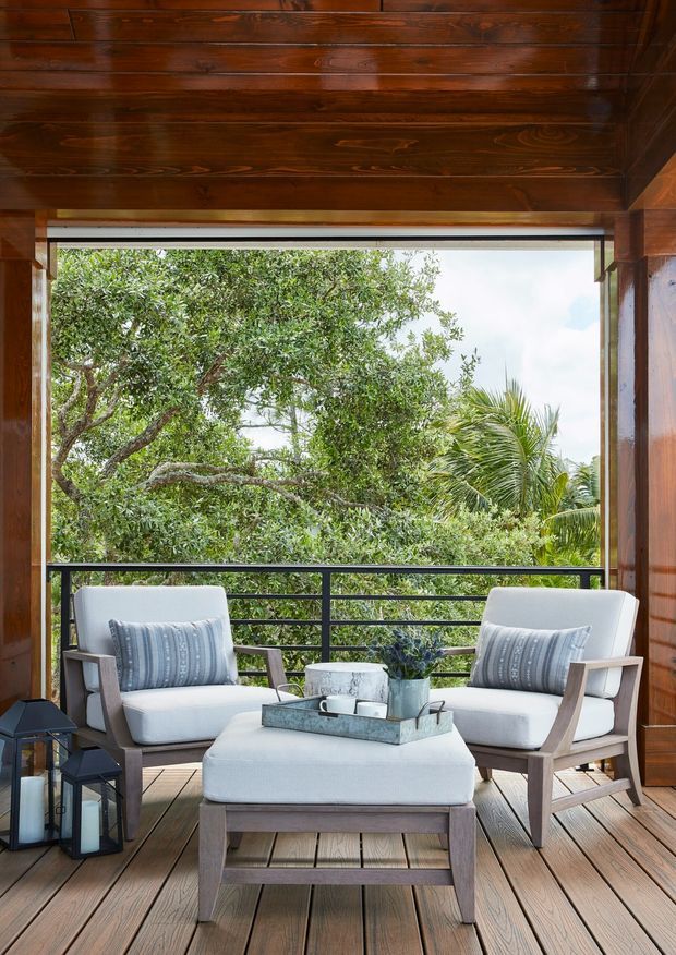 A covered outdoor terrace or balcony furnished with two light upholstered armchairs and a matching ottoman around a tray-topped coffee table. The space has warm timber wall and ceiling cladding, timber decking, dark metal railings, and lush tropical greenery beyond the open edge.