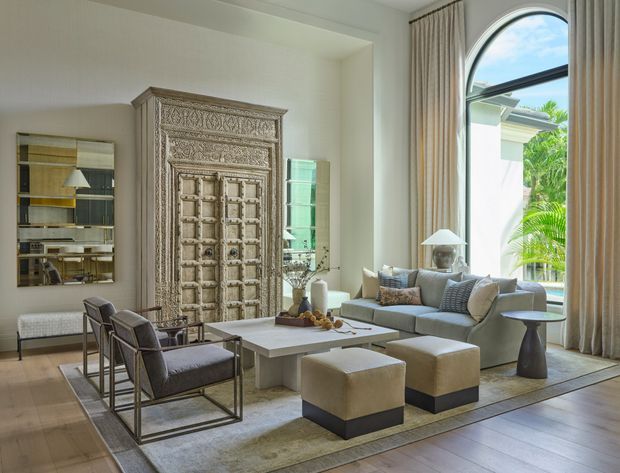 A refined, spacious living room with a very high ceiling and dramatic arched floor-to-ceiling window. The room features a pale neutral palette, large sectional sofa, upholstered ottomans, glass and metal armchairs, a low rectangular coffee table, tall curtains, a richly carved ornamental timber door or wall feature, and contemporary lighting. Tropical greenery and an exterior roofline are visible through the window.